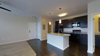 This single-floor residence offers a thoughtfully designed open-concept layout that seamlessly connects the kitchen and living areas, creating a spacious and inviting atmosphere. The kitchen is a standout feature, showcasing rich dark cabinetry that extends to the upper walls, providing ample storage space. A central kitchen island with a striking granite countertop serves as both a functional workspace and a natural gathering point, complemented by a modern sink and stylish pendant lighting overhead. Black appliances, including a refrigerator, range, and microwave, offer a sleek, cohesive aesthetic throughout the kitchen. The flooring throughout the main living area is rich, dark hardwood, adding warmth and a sense of refinement to the space. Neutral-toned walls keep the interior feeling bright and open, while recessed and flush-mount lighting ensures the space is well-lit. The entryway, visible from the main living area, features a classic paneled front door and an electrical panel access, contributing to the home's practical layout. A glimpse into an adjoining room hints at additional living spaces beyond. Overall, the residence balances modern design with functional, everyday livability.
