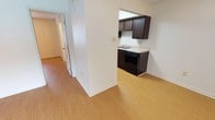 This single-floor space presents a clean, straightforward layout designed for practical everyday living. Light hardwood-style flooring runs consistently throughout, lending a cohesive and bright feel to the interior. A central structural column subtly divides the open area, creating a natural transition between the main living space and the kitchen without fully enclosing either zone.

The kitchen is compact yet functional, featuring dark espresso-toned cabinetry both above and below the counter, providing ample storage. A black appliance suite, including a range and dishwasher, is integrated neatly beneath the upper cabinets. Light-toned countertops contrast with the darker cabinetry, and a sink is positioned along the wall beneath the upper storage units. The kitchen benefits from recessed overhead lighting that keeps the space feeling open despite its modest footprint.

A hallway extends from the main area, leading toward additional rooms indicated by white panel doors. The white walls throughout maintain a neutral, clean aesthetic, reflecting available light effectively. Overall, the space offers a functional, no-frills interior with consistent finishes that create a unified and tidy environment.