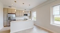 Located at 181 North Main Street in Suffolk, Virginia, this well-designed one-bedroom, one-bathroom unit offers a thoughtful layout across a single floor. The open-concept kitchen and living area serves as the heart of the space, featuring gray shaker-style cabinetry, a herringbone-patterned tile backsplash, quartz countertops, and a central island with an undermount sink. Stainless steel appliances, including a refrigerator, range, and built-in microwave, complement the modern aesthetic. Pendant lighting with gold accents hangs above the island, adding a warm, refined touch. Multiple windows throughout the kitchen and living area flood the space with natural light and offer elevated views of the surrounding treetops and Suffolk skyline. The spacious primary bedroom features notably high ceilings, creating an airy atmosphere, and includes direct access to both a dedicated closet and the full bathroom. The bathroom is appointed with white cabinetry, a warm-toned countertop, a large vanity mirror, and built-in shelving. A hallway connects the main living areas to the bedroom wing, which also includes a laundry area. The unit's consistent light-toned flooring and neutral wall palette create a cohesive, contemporary feel throughout.