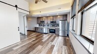 This well-appointed one-bedroom, one-bathroom unit offers a thoughtfully designed open-concept layout with contemporary finishes throughout. Wide-plank wood-look flooring flows seamlessly across the entire space, creating a cohesive and modern aesthetic.

The kitchen serves as a functional centerpiece, featuring rich, two-tone cabinetry with dark lower and medium-toned upper cabinets, a mosaic tile backsplash, stainless steel appliances including a French door refrigerator, dishwasher, range, and microwave, all framed by recessed lighting above.

The open living area is accentuated by elevated ceilings and a ceiling fan, lending a sense of spaciousness. A striking black-hardware sliding barn door adds an industrial-chic design element and provides a stylish partition between spaces.

The bedroom is a well-proportioned retreat with a large window offering urban cityscape views, filling the room with natural light. The bathroom is sleekly finished with a floating vanity, white rectangular sink, matte black fixtures, a framed mirror, and vertical LED sconce lighting.

Throughout the unit, black-framed windows frame elevated city views, connecting the interior to its vibrant urban surroundings. The overall design balances modern industrial character with clean, refined details.