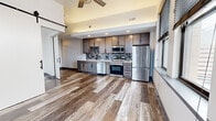 This well-appointed one-bedroom, one-bathroom unit offers a thoughtfully designed open-concept layout with contemporary finishes throughout. Wide-plank wood-look flooring flows seamlessly across the entire space, creating a cohesive and modern aesthetic.

The kitchen serves as a functional centerpiece, featuring rich, two-tone cabinetry with dark lower and medium-toned upper cabinets, a mosaic tile backsplash, stainless steel appliances including a French door refrigerator, dishwasher, range, and microwave, all framed by recessed lighting above.

The open living area is accentuated by elevated ceilings and a ceiling fan, lending a sense of spaciousness. A striking black-hardware sliding barn door adds an industrial-chic design element and provides a stylish partition between spaces.

The bedroom is a well-proportioned retreat with a large window offering urban cityscape views, filling the room with natural light. The bathroom is sleekly finished with a floating vanity, white rectangular sink, matte black fixtures, a framed mirror, and vertical LED sconce lighting.

Throughout the unit, black-framed windows frame elevated city views, connecting the interior to its vibrant urban surroundings. The overall design balances modern industrial character with clean, refined details.