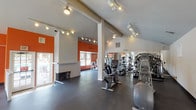Located at 43247–43265 16th St W in Lancaster, CA, this single-story fitness center is designed to provide a functional and energizing workout environment. The space features vaulted ceilings that lend an open, airy feel, with track lighting running throughout to ensure consistent illumination across the entire floor plan. Durable dark rubber flooring covers the space, offering both practicality and a clean, athletic aesthetic.

Bold orange accent walls punctuate the otherwise neutral white interior, creating a dynamic visual contrast and an energizing atmosphere reinforced by motivational text decals displayed on the walls. A notable architectural detail is the centrally positioned white fireplace, which serves as a distinctive focal point within the open layout.

The fitness center is well-stocked with a variety of equipment, including Landice treadmills, elliptical trainers, stair climbers, stationary bikes, and cable-based strength machines. Full-length mirrors along one wall enhance the sense of space and support exercise form. Large arched windows and standard windows on multiple walls allow natural light to filter in, with views of the surrounding outdoor areas. Double glass doors provide easy access to the exterior, further connecting the interior to the outdoors.