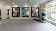 This single-floor fitness studio is thoughtfully designed to support a range of wellness activities, from yoga and stretching to cardio and strength training. The space is divided into distinct functional zones, each outfitted with purposeful equipment and finishes that contribute to a cohesive, professional atmosphere.

The primary open exercise area features neutral-toned, patterned carpet tiles that provide cushioning underfoot. A wall-mounted flat-screen television is paired with the motivational phrase "Breathe Deeply" displayed prominently above, setting a calming tone. Sanitation dispensers and a large window offering views of surrounding greenery further enhance the welcoming environment.

An adjacent zone is distinguished by a darker charcoal accent wall, which serves as a backdrop for organized fitness accessories including yoga blocks, resistance bands, and a pull-up bar rack. This area transitions into a hallway adorned with framed artwork, adding a refined touch.

The main fitness room features three large black-framed mirrors that amplify the sense of space and allow users to monitor form during workouts. A stability ball rack with assorted exercise balls and a True freestanding cardio machine round out the equipment offerings. Recessed lighting and crown molding throughout lend a polished, upscale quality to the overall space.