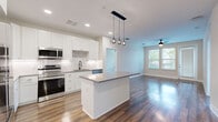 This single-floor residence showcases a contemporary design with hardwood-style flooring running throughout the entire space, lending warmth and visual continuity from room to room. The open-concept kitchen serves as a central highlight, featuring white shaker cabinetry extending to the ceiling, a marble-look tile backsplash, and a full suite of stainless steel appliances, including a range, microwave, refrigerator, and dishwasher. A kitchen island with a gray quartz-style countertop provides additional workspace and anchors the layout, while a trio of globe pendant lights adds a modern aesthetic touch overhead.

The kitchen flows seamlessly into a generously sized living area, which benefits from natural light through large windows and a glass-paneled door that leads to an outdoor balcony. A ceiling fan serves both this space and the individual bedrooms, each of which features the same hardwood-style flooring, neutral wall tones, and natural light from well-positioned windows.

The bathrooms are finished with white vanity cabinetry, gray countertops, undermount sinks, matte black hardware and fixtures, and backlit mirrors. One bathroom includes a walk-in shower with tile surround. The overall aesthetic throughout the space is clean, modern, and cohesive.
