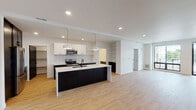 This contemporary single-floor residence showcases a thoughtfully designed open-concept layout with clean lines and a cohesive modern aesthetic. The heart of the home is a striking kitchen anchored by a large center island topped with elegant veined quartz countertops and a matte black sink. Bold dark cabinetry on the lower units contrasts sharply with crisp white upper cabinets, while a linear tile backsplash adds texture to the design. Stainless steel appliances, including a French door refrigerator, range, and microwave, complete the culinary space. Pendant lighting over the island and recessed ceiling lights provide ample illumination. A dedicated walk-in pantry offers generous storage. The kitchen opens seamlessly into a spacious living area finished with warm light-toned hardwood-style flooring that runs throughout the residence. Bedrooms are bright and airy, each featuring large windows that invite natural light and outdoor views of the surrounding neighborhood. Striking black interior doors create bold accents against the white walls across the home. The bathrooms are appointed with marble-look tile flooring, dark wood vanities, white quartz countertops, and large mirrors, blending function with refined style.