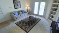 This single-floor space centers around a well-appointed living room that combines comfort and functionality. The room features wall-to-wall carpeting in a neutral tone, providing a soft and warm foundation throughout. A set of French doors with multi-pane glass panels serves as a prominent architectural feature, allowing natural light to filter into the space while offering access to the outdoors. The doors are fitted with white blinds, giving occupants control over privacy and light levels.

The living area is furnished with a generously sized, cushioned sofa accented with blue throw pillows, positioned to take advantage of both the natural light and the room's layout. A glass-top coffee table with an ornate wrought-iron base sits atop a dark, patterned area rug featuring an elegant leafy border design, adding visual interest and defining the seating zone.

Additional details include a tall floor lamp and a decorative wrought-iron candle stand that contribute to the room's ambient lighting. A framed piece of artwork adorns the wall above the sofa, adding a touch of character. Overall, the space presents a tidy, inviting atmosphere with a classic, understated aesthetic.