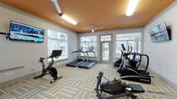 Located at 230 Pine Knot Lane in Graham, NC, this fitness center is a thoughtfully designed, single-floor facility offering a comprehensive workout environment. The space is defined by its striking warm-orange ceiling accented with ceiling fans and fluorescent lighting, creating an energetic yet comfortable atmosphere throughout.

The cardio zone features multiple treadmills with individual screens, stationary bikes including a Cascade rowing machine, an elliptical trainer, and a spin cycle — all positioned near large windows that allow ample natural light and views of the surrounding community. Glass doors provide direct outdoor access to what appears to be a pool area, enhancing the sense of openness.

The strength training area is equally well-appointed, housing a multi-station cable machine, a full rack of dumbbells, free weights, and a dedicated functional training zone complete with a punching bag and TRX suspension system. A wall-mounted mirror and flat-screen TV further enhance the training experience.

Additional amenities include a water fountain station with a hydration refill option, wall-mounted TVs, and organized storage for fitness accessories such as stability balls and resistance tools. The patterned carpet tile flooring ties the space together, offering both durability and visual appeal throughout this fully equipped fitness facility.