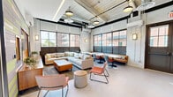Located at 321 Ellis St in New Britain, Connecticut, this single-floor amenity space blends industrial architecture with contemporary design to create a vibrant, multifunctional communal environment. The layout is divided into two distinct zones that flow seamlessly together.

The main lounge area is defined by soaring ceilings with exposed ductwork, painted brick walls, and a wall of large, floor-to-ceiling industrial-style windows that flood the space with natural light and offer views of the surrounding urban streetscape. A generous sectional sofa anchors a comfortable seating arrangement, complemented by accent chairs in warm orange tones and a variety of casual side tables. A standout feature wall combines reclaimed wood panels with colorful horizontal accents in green and gray, framing a wall-mounted television and built-in media console. Adjacent dining tables with modern chairs provide additional flexible seating, and a kitchenette with bar seating and stainless steel appliances adds convenience.

The second zone serves as a dedicated game room, featuring a full-size pool table set beneath oversized pendant lights. Wainscoted walls in cool gray, a wall-mounted television, and a cue rack complete the space. Large glass-paneled doors connect both areas, maintaining an open, airy feel throughout. The polished concrete floors and pendant lighting tie the entire space together with an inviting, modern-industrial aesthetic.