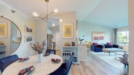 This single-floor residence combines contemporary design with vibrant, curated interiors across a thoughtfully planned open-concept layout. Light wood flooring flows through the main living areas, connecting a dining space, living room, and kitchen in a seamless arrangement. The dining area features a marble-top round table beneath a sculptural brass chandelier, set against a soft watercolor accent wall with a round gold-framed mirror as a focal point. A built-in bar cart and partial dividing wall elegantly separate the dining zone from the kitchen, which is equipped with stainless steel appliances. The living room is anchored by a sliding glass door that opens to a private balcony overlooking a serene lakefront, filling the space with natural light and tranquil views. The primary bedroom is a standout space, featuring a bold botanical wallpaper accent wall, plush carpeting, generous natural light from a large window, and access to a spacious walk-in closet with built-in shelving. The bathroom is clean and bright, finished with wood-look flooring, a white vanity, and a glass-enclosed walk-in shower. Throughout, the residence strikes a balance between eclectic personality and functional, well-organized living.