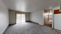 This single-floor residence offers a well-organized layout featuring three bedrooms, two bathrooms, a living room, a hallway, and a dedicated closet. The living room is a generous, open space finished with neutral-toned carpet and accented by warm wood trim baseboards. A full-width sliding glass door with vertical blinds fills the room with natural light and provides direct access to an outdoor balcony area. A wall-mounted air conditioning unit serves the living space alongside baseboard heating units found throughout the home.

The primary bedroom is notably spacious, offering ample room for furnishings, and includes a built-in closet with sliding wood-trimmed doors. The two additional bedrooms are similarly carpeted and include closet storage, with one bedroom also featuring a window with a view to the exterior. Each bedroom benefits from baseboard heating for year-round comfort.

The two bathrooms offer functional designs. One bathroom features a full tub and shower combination with classic beige square tile surrounds and chrome fixtures. The second bathroom includes a wood-cabinet vanity with a single-basin sink, a large mirror with overhead lighting, and wood-look vinyl plank flooring. The consistent use of warm wood-toned trim and cabinetry throughout ties the interior together with a cohesive, understated aesthetic.