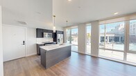 This single-floor unit showcases a contemporary design with an open-concept layout that seamlessly connects the kitchen, dining, and living areas. Light hardwood flooring flows consistently throughout, complementing the crisp white walls and clean architectural lines.

The kitchen is a standout feature, centered around a large island with a white quartz countertop, dark wood cabinetry, and a gold-toned faucet with an undermount sink. Pendant lighting hangs above the island, adding warmth to the space. The full suite of stainless steel appliances includes a refrigerator, microwave, and range, all framed by a white tile backsplash and dark upper and lower cabinetry.

Floor-to-ceiling windows line the living area, flooding the space with abundant natural light and offering open urban street views. The living area is generously proportioned and flows naturally from the kitchen.

The bedroom includes a large floor-to-ceiling window with city views and direct access to a private bathroom. The bathroom features hexagonal tile flooring, a floating vanity, a large mirror, and modern black and gold fixtures, creating a clean, refined aesthetic throughout.