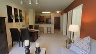 This single-floor residence offers an open-concept layout that thoughtfully integrates the living, dining, and kitchen areas into one cohesive and functional space. The living area features plush carpeting and is bathed in warm ambient lighting, creating a welcoming atmosphere complemented by tasteful décor including a cushioned sofa and accent lamp. A bold, dark accent wall anchors the space and adds a striking visual contrast to the neutral tones throughout.

The dining area is positioned adjacent to the living space and features a dark-finished dining table with upholstered chairs, adding a touch of elegance to everyday meals. Decorative candle holders on the table contribute to the warm, inviting ambiance.

The kitchen is designed with functionality in mind, boasting rich dark wood cabinetry, granite countertops, and black appliances including a refrigerator. A raised breakfast bar with bar stools provides an additional casual dining or entertaining option, creating a natural divide between the kitchen and the open living space.

Recessed lighting and overhead fixtures throughout ensure the space is well-lit, while the clean, neutral color palette of the walls and flooring creates a sense of openness. A hallway near the entry leads to additional areas of the home, completing this thoughtfully designed and versatile layout.