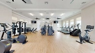 Located at 200 Villas Dr in Lavon, Texas, this well-appointed fitness center offers a generously sized, single-floor workout facility designed to support a full range of exercise routines. The space features light hardwood-style flooring throughout, crisp white walls, and multiple large windows that flood the interior with natural light while providing views of the surrounding community and pool area. Multiple oversized ceiling fans are mounted throughout, ensuring ample air circulation during workouts.

The cardio zone is equipped with a row of modern treadmills, a stair climber, a rowing machine, and a stationary bike, all positioned along the windowed walls to maximize natural light. The strength training area is equally comprehensive, featuring multi-station cable machines, a power rack, a dumbbell rack with free weights, kettlebells, medicine balls, and adjustable benches. Wall-mounted mirrors in the free weights section enhance the sense of space and assist with form. Mounted flat-screen TVs are positioned for easy viewing across multiple workout zones.

A dedicated fitness studio area with glass-door entry provides additional space for group or individual training. A water fountain is conveniently located near the entrance. The overall ambiance is clean, modern, and energizing, making this facility a standout amenity for the Villas community.