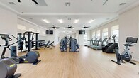 Located at 200 Villas Dr in Lavon, Texas, this well-appointed fitness center offers a generously sized, single-floor workout facility designed to support a full range of exercise routines. The space features light hardwood-style flooring throughout, crisp white walls, and multiple large windows that flood the interior with natural light while providing views of the surrounding community and pool area. Multiple oversized ceiling fans are mounted throughout, ensuring ample air circulation during workouts.

The cardio zone is equipped with a row of modern treadmills, a stair climber, a rowing machine, and a stationary bike, all positioned along the windowed walls to maximize natural light. The strength training area is equally comprehensive, featuring multi-station cable machines, a power rack, a dumbbell rack with free weights, kettlebells, medicine balls, and adjustable benches. Wall-mounted mirrors in the free weights section enhance the sense of space and assist with form. Mounted flat-screen TVs are positioned for easy viewing across multiple workout zones.

A dedicated fitness studio area with glass-door entry provides additional space for group or individual training. A water fountain is conveniently located near the entrance. The overall ambiance is clean, modern, and energizing, making this facility a standout amenity for the Villas community.