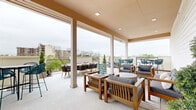 This rooftop terrace is a well-appointed outdoor amenity space designed for both relaxation and social gatherings. The terrace is divided into distinct functional zones, seamlessly blending covered and open-air areas. Sturdy white columns support a warm-toned overhead structure fitted with recessed lighting, providing shade and ambiance for the covered lounge section. The flooring throughout is a clean, light-toned surface that contributes to the bright, airy feel of the space.

The covered lounge area features craftsman-style wooden furniture with plush gray cushions, arranged around a fluted cylindrical coffee table, creating an inviting setting for conversation. A large-format connect-four game adds a playful, social element to the space. Moving outward, the open-air section offers additional seating with teal woven chairs and dark bar-height tables positioned along the railing, taking full advantage of the panoramic views of the city skyline and surrounding tree canopy.

A built-in wet bar station with a sink and counter space is conveniently located near the transition between covered and open areas, supporting outdoor entertaining. Potted topiary accents and area rugs add refined finishing touches throughout. The iron railing perimeter frames sweeping views, making this rooftop terrace an elevated and versatile outdoor retreat.