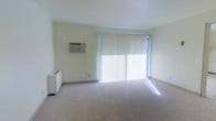 This single-floor space offers a clean, minimalist layout designed for comfortable everyday living. The main living area is finished with soft, neutral-toned wall-to-wall carpeting that adds warmth underfoot, while the smooth white walls and white ceiling contribute to a bright, airy atmosphere throughout.

A prominent sliding glass door serves as a key architectural feature, framed by floor-to-ceiling vertical blinds that allow for easy light control. When the blinds are open, natural light streams generously into the room, enhancing the sense of openness and space. A wall-mounted air conditioning unit is conveniently positioned on one wall, ensuring climate comfort year-round, and is complemented by a baseboard heating unit tucked into the corner, providing additional temperature control options.

The overall ambiance of the space is calm and neutral, offering a versatile blank canvas well-suited for a variety of furniture arrangements and personal styles. The uncluttered layout and abundance of natural light make the interior feel welcoming and functional, while the simple, well-maintained finishes reflect a practical and livable design throughout.