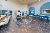Located at 301 High Tower Blvd in Pittsburgh, PA, this expansive single-floor amenity space is designed with a modern, upscale aesthetic that blends functionality with refined style. The grand entrance lobby commands immediate attention with soaring double-height ceilings, floor-to-ceiling windows that flood the space with natural light, dramatic pendant and chandelier lighting, and richly veined dark marble flooring. A sleek reception counter with a white quartz top and navy tile base anchors the welcoming area. The main living lounge features generous seating arrangements with plush gray and cognac leather furniture, a bold blue accent wall with arched detailing, and textured wallcovering that adds visual depth. A second lounge area showcases a wall-mounted fireplace, large-format artwork, and expansive windows offering outdoor views, creating a warm yet contemporary retreat. The kitchen and dining area is outfitted with dark cabinetry, gold hardware, white quartz countertops, open metal shelving, and a large island—ideal for entertaining. A billiards table adds a recreational touch to the adjacent gathering space. Two conference rooms provide polished settings for meetings, featuring statement wallcoverings, mounted displays, stylish pendant lighting, and open shelving. Connecting hallways tie the entire space together with a seamless, cohesive flow.