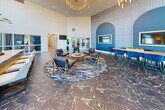 Located at 301 High Tower Blvd in Pittsburgh, PA, this expansive single-floor amenity space is designed with a modern, upscale aesthetic that blends functionality with refined style. The grand entrance lobby commands immediate attention with soaring double-height ceilings, floor-to-ceiling windows that flood the space with natural light, dramatic pendant and chandelier lighting, and richly veined dark marble flooring. A sleek reception counter with a white quartz top and navy tile base anchors the welcoming area. The main living lounge features generous seating arrangements with plush gray and cognac leather furniture, a bold blue accent wall with arched detailing, and textured wallcovering that adds visual depth. A second lounge area showcases a wall-mounted fireplace, large-format artwork, and expansive windows offering outdoor views, creating a warm yet contemporary retreat. The kitchen and dining area is outfitted with dark cabinetry, gold hardware, white quartz countertops, open metal shelving, and a large island—ideal for entertaining. A billiards table adds a recreational touch to the adjacent gathering space. Two conference rooms provide polished settings for meetings, featuring statement wallcoverings, mounted displays, stylish pendant lighting, and open shelving. Connecting hallways tie the entire space together with a seamless, cohesive flow.