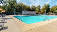 This outdoor pool area offers a well-appointed recreational retreat designed for relaxation and leisure. At its center is a generously sized rectangular swimming pool filled with clear, blue-tinted water, accessible via stainless steel handrails. The surrounding concrete sun deck provides ample space for lounging, outfitted with numerous adjustable chaise lounge chairs arranged along the poolside for optimal sun exposure. Multiple seating areas are scattered throughout the deck, each featuring round metal tables paired with chairs and shaded by vibrant orange umbrellas, ideal for outdoor dining or casual gatherings. A compact utility structure, finished with white siding and a shingled roof, sits at one end of the pool area and provides additional support facilities. The space is fully enclosed by a decorative wrought iron fence, ensuring both security and an open, airy feel. A standout feature of this pool area is its scenic backdrop — one side of the enclosure opens to a tranquil waterfront view, with a pond and open green space visible beyond the fence, enhancing the overall outdoor ambiance. Mature trees lining the perimeter provide natural shade and a lush, green setting under open skies, making this an inviting space for outdoor recreation.