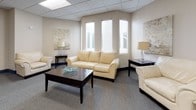 This single-floor space is thoughtfully designed to offer both comfort and practicality, making it well-suited for professional or community use. The layout is open and accommodating, with distinct zones that serve different purposes while maintaining a cohesive aesthetic throughout.

The main lounge area is furnished with cream leather sofas and matching armchairs arranged around a dark wood coffee table, creating an inviting and relaxed atmosphere. Tall windows allow natural light to filter in, brightening the neutral-toned walls and grey carpeted floors. Decorative wall art and table lamps add warmth and a polished finish to the space.

A separate seating nook features a single cream leather armchair paired with a side table and lamp, offering a quieter, more private corner ideal for individual use or one-on-one conversations.

The meeting and dining area includes a dark wood rectangular table surrounded by matching chairs, providing a functional space for group gatherings or discussions. Interior windows with views into adjacent rooms and a clearly marked emergency exit door reflect the space's attention to safety and accessibility. Blue baseboards provide a subtle accent throughout, tying the rooms together visually.

Overall, the space presents a clean, professional environment with a welcoming ambiance, suitable for a range of uses including meetings, consultations, or community gatherings.