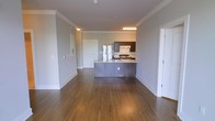 This single-floor space is designed with an open-concept layout that seamlessly integrates the living area and kitchen. Rich hardwood flooring extends throughout, adding warmth and continuity to the overall aesthetic. Crown molding lines the ceiling, lending a polished, refined finish to the neutral gray walls.

The kitchen serves as a functional focal point, anchored by a peninsula-style island with a stainless steel countertop and dark cabinetry below, offering both prep space and a natural divider between the kitchen and living areas. Dark upper cabinetry complements the lower units, creating a cohesive design. Stainless steel appliances, including a refrigerator and a range with an overhead ventilation hood, complete the kitchen setup.

The living area is generously proportioned, with clean, unobstructed sightlines that emphasize the openness of the floor plan. Recessed and flush-mount ceiling lighting provides even illumination throughout the space. Multiple doorways lead to additional rooms, suggesting a well-connected layout. The combination of neutral tones, hardwood floors, and classic architectural details gives the space a timeless, move-in-ready appeal.