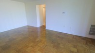 This single-floor space presents a straightforward and functional layout designed to accommodate a variety of living arrangements. The main living area is immediately defined by its classic parquet wood flooring, which features a warm, honey-toned finish with a distinctive block pattern that adds visual texture and timeless character to the room. The walls are finished in a clean, neutral white, creating a bright and airy atmosphere that allows natural light to reflect throughout the space. The open nature of the main room provides a generous sense of space, offering flexibility in how the area can be arranged and utilized. A doorway opening off the main room leads to an adjacent hallway, providing access to the remaining areas of the unit. The transition between rooms is clean and unobstructed, contributing to a smooth flow throughout the floor plan. Electrical outlets and light switches are positioned at standard intervals along the walls, reflecting a practical and well-considered layout. Overall, the space offers a neutral, move-in-ready canvas with durable flooring and ample wall space, making it well-suited for a range of furnishing styles and functional uses.