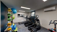 Located at 3063 Sprowl Rd in Louisville, Kentucky, this single-floor fitness center is designed to support a well-rounded workout experience. The space is organized to accommodate both cardiovascular training and strength work, with distinct zones dedicated to each discipline.

The cardio area is equipped with multiple TRUE brand treadmills and elliptical machines, each featuring integrated display screens for performance tracking. A spin bike provides an additional low-impact cycling option. The flooring throughout consists of patterned blue and grey carpet tiles that define the space and provide a degree of cushioning underfoot.

The strength training area includes a two-tier dumbbell rack stocked with a range of free weights, accompanied by an adjustable workout bench. A wall-mounted flat-screen television overlooks this zone, and a piece of abstract artwork adds a decorative accent to the otherwise functional space.

Recessed LED panel lighting illuminates the room evenly, while a wall-mounted mini-split air conditioning unit provides climate control. Natural light enters through windows on the teal-accented accent wall, contributing a sense of openness. A large wall mirror near the cardio equipment enhances the perception of space. Exercise accessories, including stability balls and yoga mats, round out the available amenities.