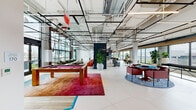 This impressive single-floor space is designed to foster social interaction and comfort, combining industrial-chic architecture with vibrant, thoughtfully curated interiors. The open-plan layout is anchored by soaring ceilings with exposed black steel framework, structural beams, and an industrial-style grid that adds a bold, contemporary character throughout.

The main lounge and recreation area features a full-size pool table set atop a richly colored area rug, alongside multiple seating zones furnished with plush sofas and lounge chairs. A dedicated games nook includes a poker-style table surrounded by upholstered seating, positioned on a decorative circular rug. Throughout the space, large potted tropical plants introduce vibrant greenery and a sense of vitality.

The kitchen area is a standout feature, showcasing a dramatic floor-to-ceiling backsplash composed of shimmering teal mosaic tiles, complemented by sleek dark cabinetry and a bar-height counter with contemporary stools. An adjoining open dining area provides a long communal table bathed in natural light from wraparound floor-to-ceiling windows.

Several lounge nooks are positioned directly along the glass curtain walls, offering sweeping panoramic views of the surrounding cityscape and direct access to a landscaped outdoor terrace. The large-format hexagonal tile flooring, wood ceiling accents, and layered lighting complete this dynamic, amenity-rich environment.
