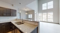 Located at 17016 East Indiana Parkway in Spokane Valley, WA, this contemporary single-floor unit impresses with its dramatic vaulted ceilings and open, airy layout. The open-concept kitchen is thoughtfully designed with rich dark cabinetry, granite countertops, a classic white subway tile backsplash, and a full suite of stainless steel appliances including a refrigerator, range, microwave, and dishwasher. Pendant lighting above the kitchen peninsula adds a stylish touch, while light hardwood-style flooring runs throughout the main living areas. The spacious living room is a standout feature, boasting soaring two-story ceilings and a generous wall of windows that flood the space with natural light and offer views of the surrounding neighborhood. A private entrance with adjacent closet storage provides a welcoming foyer area. The bedroom continues the vaulted ceiling design, with a ceiling fan and a window framing outdoor views. The bathroom is clean and functional, featuring a soaking tub and light flooring. A dedicated in-unit laundry area with a full-size washer and dryer adds everyday convenience. The unit's neutral palette and modern finishes create a bright, polished ambiance throughout.