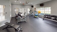 Located at 9079 SR-5 in Douglasville, Georgia, this fitness center offers a well-appointed, single-floor layout designed to accommodate a comprehensive range of workout needs. The space is finished with durable rubber flooring throughout, providing a safe and practical surface suited to both cardio and strength training activities.

The facility is organized into distinct workout zones. A dedicated cardio area houses treadmills, ellipticals, stationary bikes, and cycling bikes, all positioned near large, multi-pane windows that allow ample natural light and views of the surrounding exterior. A strength training zone features multi-function cable machines, weight benches, and a dumbbell rack, giving users access to versatile resistance training options. Resistance bands and stability balls are also available for functional fitness exercises.

Architecturally, the space features crown molding, white-trimmed columns, and gray-painted walls that lend a clean, polished appearance. Recessed lighting and ceiling fans provide consistent illumination and air circulation throughout. Large wall mirrors in one section visually expand the space and assist users during workouts. Wall-mounted televisions offer entertainment options. French doors and generously sized windows connect the interior to natural light, contributing to an open and motivating workout environment.