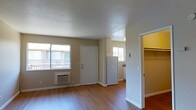 This single-floor unit offers a straightforward, functional layout designed for comfortable everyday living. The main living area is finished with warm hardwood-style flooring that extends throughout, complementing the neutral taupe-toned walls and crisp white baseboards and door trim. The textured ceiling adds a subtle architectural detail overhead.

Two windows in the main living space allow natural light to filter in, offering views of an adjacent outdoor balcony area. A wall-mounted air conditioning unit is positioned beneath one of the windows, providing convenient climate control. The front entry door opens directly into this living space, creating an accessible and open feel.

Adjacent to the living area, a compact kitchen is visible, featuring white cabinetry and a refrigerator. The kitchen connects seamlessly to the main space without visual barriers. A doorway leads to an additional room that includes a built-in closet with overhead shelving, offering practical storage. A separate hallway visible in the interior provides access to additional areas of the unit. Overall, the space presents a clean, move-in-ready environment with a cohesive, neutral palette throughout.
