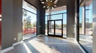This striking single-floor commercial space makes an immediate impression with its soaring double-height ceilings and expansive floor-to-ceiling windows that flood the interior with abundant natural light. The entry foyer features sleek hardwood-style flooring, dark-framed glass double doors, and a dramatic sputnik chandelier, setting an upscale tone from the moment of arrival. Bold red accent columns and charcoal gray walls create a confident, contemporary color palette that carries throughout the space. The main lounge area is generously proportioned, furnished with plush navy blue velvet seating arrangements, gold-legged accent chairs, and layered area rugs that define conversation areas beneath additional statement chandeliers. A branded feature wall with neon signage anchors one end of the lounge, adding an energetic focal point. A dedicated workspace or leasing station features a vivid red countertop, dark velvet chairs with gold legs, shiplap wall cladding, and red pendant lighting above, complemented by framed mirrors and lush tropical greenery. Private office areas with red accent walls and dark wood desks provide functional workspace. Throughout the space, large-format artwork, carefully curated decor, and an abundance of natural light from oversized windows contribute to a polished, welcoming, and professionally designed environment.