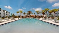 Located at 100 Windflower Ln in Daytona Beach, Florida, this outdoor amenity area offers a resort-inspired experience designed for relaxation and recreation. At the heart of the space is a generously sized swimming pool with crystal-clear blue water, flanked by a wide, well-appointed sun deck lined with lounge chairs and teal-accented umbrellas. Towering palm trees are thoughtfully placed throughout the deck, providing both shade and a tropical aesthetic that complements the sunny Florida climate.

Surrounding the pool, lush green lawn areas serve as dedicated spaces for outdoor activities, including lawn games such as cornhole and ping pong, making the area ideal for social gatherings. Hanging egg chairs and additional seating nodes are interspersed among the palms, offering more intimate lounging options.

A beautifully designed single-story clubhouse pavilion anchors one end of the pool area, featuring a covered columned veranda with large glass doors that open to the pool deck, creating a seamless indoor-outdoor connection. The structure's white exterior with a charcoal roof complements the clean, coastal architectural style of the broader community. Black iron fencing encloses the perimeter, providing security while maintaining open sightlines to the surrounding landscaping and multi-story residential buildings beyond.