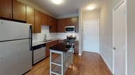 This single-floor unit showcases a clean, contemporary design with warm hardwood-style flooring flowing throughout the main living areas. The open-concept kitchen is well-equipped with rich medium-toned wood cabinetry, white subway tile backsplash, granite-style countertops, and a full suite of stainless steel appliances including a refrigerator, gas range, microwave, and dishwasher. A freestanding kitchen island provides additional prep and storage space, and the layout connects seamlessly to the entry foyer and main living area.

The spacious living area is bathed in natural light from a large sliding glass door that opens onto a private balcony, offering views of the surrounding neighborhood. The neutral gray walls and hardwood-style flooring create a bright, airy atmosphere throughout.

The bedroom features plush carpeting, a ceiling fan with light fixture, and dual windows that allow for generous natural light. The bathroom is accessed via a short hallway and features a generous wood vanity with dark countertop, an integrated sink, a large mirror with overhead lighting, and hardwood-style flooring that continues the cohesive aesthetic of the unit. The overall design emphasizes functionality and modern comfort throughout.