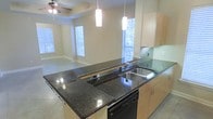 This single-story residence showcases an open-concept layout that seamlessly connects the kitchen and living areas, creating a spacious and cohesive environment. The kitchen is a standout feature, anchored by a large island with rich, dark granite countertops that offer generous prep and entertainment space. Light maple cabinetry lines the walls and island base, providing ample storage while contributing a warm, contemporary aesthetic. A stainless steel sink is integrated into the island, complemented by a sleek faucet and a built-in dishwasher beneath the counter. Two modern rectangular pendant lights hang above the island, adding focused task lighting and a polished design touch. The kitchen connects naturally to the living area, which features tile flooring throughout and a ceiling fan overhead. Multiple windows dressed with blinds allow abundant natural light to fill both spaces, enhancing the open and airy atmosphere. The neutral wall tones and clean architectural lines throughout contribute to a bright, well-balanced interior that is both functional and inviting.