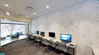 This single-floor business center offers a well-equipped and thoughtfully designed workspace for productivity. A long granite-topped counter runs along one wall, accommodating a row of desktop computers paired with ergonomic mesh-back chairs, providing ample workstation space for multiple users simultaneously. The countertop's light stone finish adds a polished, professional aesthetic to the room.

The standout design element is the expansive decorative wall featuring a soft white background adorned with scattered blue watercolor-style bubble motifs, lending the space a calm, contemporary feel. Recessed lighting in the drop ceiling ensures consistent, even illumination throughout the room. The flooring consists of patterned commercial-grade carpeting in dark tones, offering a practical and durable foundation that complements the room's overall color palette.

A glass entryway is visible at one end of the room, allowing natural light to filter in and contributing to the open, accessible atmosphere of the space. The overall ambiance is clean, modern, and functional, making it well-suited for business and professional use.