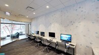 This single-floor business center offers a well-equipped and thoughtfully designed workspace for productivity. A long granite-topped counter runs along one wall, accommodating a row of desktop computers paired with ergonomic mesh-back chairs, providing ample workstation space for multiple users simultaneously. The countertop's light stone finish adds a polished, professional aesthetic to the room.

The standout design element is the expansive decorative wall featuring a soft white background adorned with scattered blue watercolor-style bubble motifs, lending the space a calm, contemporary feel. Recessed lighting in the drop ceiling ensures consistent, even illumination throughout the room. The flooring consists of patterned commercial-grade carpeting in dark tones, offering a practical and durable foundation that complements the room's overall color palette.

A glass entryway is visible at one end of the room, allowing natural light to filter in and contributing to the open, accessible atmosphere of the space. The overall ambiance is clean, modern, and functional, making it well-suited for business and professional use.