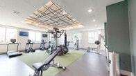 This fitness center offers a thoughtfully designed workout facility across a single, open floor plan. The space is immediately distinguished by its eye-catching coffered ceiling featuring a bold geometric grid pattern with recessed lighting, creating a visually dynamic overhead focal point that elevates the overall aesthetic.

The layout is organized to accommodate a wide range of fitness activities. A central green turf zone serves as the functional training hub, hosting a multi-station cable machine, a heavy punching bag, TRX suspension trainers, medicine balls, and an adjustable weight bench. Along the perimeter, a dedicated cardio row includes treadmills, elliptical trainers, and stationary bikes from brands such as TRUE Fitness. Additional strength equipment, including a leg press/curl machine and a cable weight stack, is positioned throughout the space.

A full dumbbell rack with mirror backing occupies one wall, providing ample free weight options. The flooring combines dark rubberized material with the central turf section, offering both durability and versatility.

Natural light pours in through multiple large windows along the walls, offering views of the surrounding exterior. An accent wall in deep green, paired with contemporary artwork, adds a refined design touch. A glass-door entry connects the fitness center to adjacent building amenities, completing this comprehensive and well-appointed workout environment.