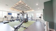 This fitness center offers a thoughtfully designed workout facility across a single, open floor plan. The space is immediately distinguished by its eye-catching coffered ceiling featuring a bold geometric grid pattern with recessed lighting, creating a visually dynamic overhead focal point that elevates the overall aesthetic.

The layout is organized to accommodate a wide range of fitness activities. A central green turf zone serves as the functional training hub, hosting a multi-station cable machine, a heavy punching bag, TRX suspension trainers, medicine balls, and an adjustable weight bench. Along the perimeter, a dedicated cardio row includes treadmills, elliptical trainers, and stationary bikes from brands such as TRUE Fitness. Additional strength equipment, including a leg press/curl machine and a cable weight stack, is positioned throughout the space.

A full dumbbell rack with mirror backing occupies one wall, providing ample free weight options. The flooring combines dark rubberized material with the central turf section, offering both durability and versatility.

Natural light pours in through multiple large windows along the walls, offering views of the surrounding exterior. An accent wall in deep green, paired with contemporary artwork, adds a refined design touch. A glass-door entry connects the fitness center to adjacent building amenities, completing this comprehensive and well-appointed workout environment.