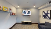 This single-floor space is designed as a comfortable lounge and entertainment area. The room features neutral gray walls paired with dark baseboards, creating a clean and modern aesthetic. Recessed ceiling lights provide even, functional illumination throughout the space. A large flat-screen television is prominently wall-mounted at the center, serving as the focal point of the room, with a low-profile media console positioned beneath it for storage and display. To one side, a wall-mounted shelf holds a selection of board games and activities, adding a recreational element to the space. A textured area rug anchors the seating zone, and a dark upholstered chair is visible at the edge of the room, contributing to the relaxed atmosphere. A decorative branch arrangement in a floor vase adds a subtle organic touch to the corner. Overall, the space is thoughtfully arranged for leisure and social gathering, with a practical layout that balances entertainment amenities with understated, contemporary design elements.