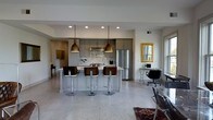 Located at 721 N High St in Columbus, Ohio's vibrant Short North Arts District, this single-floor space offers a thoughtfully designed open-concept layout that seamlessly integrates living, dining, and kitchen areas. The kitchen is a standout feature, showcasing gray cabinetry, a decorative tile backsplash, stainless steel appliances including a full-size refrigerator, and a spacious kitchen island topped with a light countertop. Three stylish bar stools with wood and chrome accents line the island, complemented overhead by a pair of warm brass pendant lights that add an industrial-chic touch. Polished terrazzo flooring extends throughout the space, lending a clean and sophisticated character to the interior. Recessed lighting in the ceiling provides even illumination across the open floor plan. A dedicated workspace area near a natural-light-filled window offers a functional nook complete with a desk and office chair. A framed gold mirror adorns the wall nearby, adding a classic accent to the modern surroundings. The overall ambiance balances contemporary design with warm, inviting tones, making this space both practical and visually refined — ideal for those who appreciate modern urban living in one of Columbus's most sought-after neighborhoods.