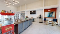 Located at 4800 N Star Ave in Panama City, Florida, this single-story community clubhouse delivers a polished and inviting atmosphere designed for both relaxation and social engagement. The space is anchored by a dramatic, voluminous great room with soaring ceilings that amplify the sense of openness and airiness throughout. Large windows along the front facade flood the interior with natural light, complementing the clean white walls and expansive light-toned tile flooring.

At the heart of the main lounge sits a substantial textured blue-grey credenza with brass accents, serving as a stylish focal point. Comfortable seating arrangements are thoughtfully positioned nearby, while a striking cascading ring chandelier overhead adds a sophisticated design element. An accent wall in warm terracotta anchors one corner of the room, displaying large-scale abstract artwork and a built-in bench with cushioned seating.

The kitchenette area is well-appointed with granite countertops, white cabinetry, a beverage refrigerator, stainless steel appliances, open shelving, and a wall-mounted flat-screen television. A classic red popcorn cart adds a playful, welcoming touch near the entrance. An adjacent workspace area features a desk and seating for focused tasks. The overall ambiance is contemporary, bright, and community-oriented.