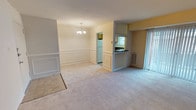 This single-floor residence offers a comfortable and functional layout designed to maximize living space. The main living and dining area is unified by soft, neutral-toned carpeting that runs throughout, creating a warm and cohesive feel underfoot. Classic wainscoting panels line the lower walls of the dining area, adding a touch of traditional architectural character, while a modest chandelier overhead provides a defined focal point for the dining space.

The living area transitions naturally toward a semi-open kitchen, separated by a half-wall breakfast bar with a dark countertop. The kitchen features tile flooring, providing a practical distinction between the cooking and living zones. A window in the kitchen allows for natural light to filter into the space, and a full-size refrigerator is visible, indicating a well-equipped cooking area.

Natural light enters the main living space through a sliding glass door or large window along one wall, fitted with vertical blinds that allow for adjustable brightness. The overall color palette is light and neutral — creamy whites and soft yellows — contributing to an airy and inviting ambiance throughout the home. The single-floor layout ensures easy accessibility and a straightforward flow between the living, dining, and kitchen areas.