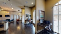 This single-floor community clubhouse is thoughtfully designed to offer residents a variety of amenity spaces, all unified by rich, dark hardwood flooring and warm neutral tones. The welcoming lounge area features a comfortable seating arrangement with a plush gold sofa, teal accent chairs, and an area rug, complemented by decorative framed artwork and a wall-mounted flat-screen television beneath a statement pendant light fixture. The business center provides multiple dedicated workstations with wall-mounted monitors, ergonomic seating, and natural light streaming through arched windows dressed with horizontal blinds. A casual dining and social area showcases tall bistro tables and bar-height seating positioned beside large arched windows with flowing striped drapery, offering views of the surrounding greenery. The entertainment room features a classic arcade game cabinet, large-screen television, and sleek modern seating, accented by bold orange walls for a vibrant atmosphere. A fully equipped kitchenette offers ample cabinetry, dark granite countertops, a tile backsplash, and a refrigerator, ideal for catering and social events. A game room with a pool table rounds out the amenities, visible through the open-concept corridor. Throughout, high ceilings, recessed lighting, and decorative pendant fixtures enhance the spacious, inviting ambiance of this well-rounded community facility.