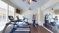 This dedicated fitness room is thoughtfully designed to support a full range of exercise activities within a comfortable, single-floor layout. The space features rich, dark hardwood-style flooring that runs throughout, providing a clean and durable surface suited for high-traffic use. Two ceiling fans with light fixtures are positioned overhead, ensuring consistent air circulation and a well-lit environment during workouts.

A row of True-brand cardio equipment, including treadmills and an elliptical trainer, lines one side of the room, offering multiple options for cardiovascular exercise. Adjacent to the cardio machines stands a multi-cable strength training station, providing versatility for resistance-based workouts. A large floor-to-ceiling mirror spans one wall, enhancing the sense of space while allowing users to monitor their form during exercise.

Natural light filters in through windows fitted with blinds, contributing to a bright and welcoming atmosphere. A wall-mounted flat-screen television is positioned for easy viewing from the cardio equipment, adding an entertainment element to workouts. Crown molding along the ceiling adds a subtle architectural detail, elevating the overall finish of the space. The room is well-ventilated with visible ceiling vents, ensuring comfort during extended exercise sessions.