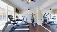 This dedicated fitness room is thoughtfully designed to support a full range of exercise activities within a comfortable, single-floor layout. The space features rich, dark hardwood-style flooring that runs throughout, providing a clean and durable surface suited for high-traffic use. Two ceiling fans with light fixtures are positioned overhead, ensuring consistent air circulation and a well-lit environment during workouts.

A row of True-brand cardio equipment, including treadmills and an elliptical trainer, lines one side of the room, offering multiple options for cardiovascular exercise. Adjacent to the cardio machines stands a multi-cable strength training station, providing versatility for resistance-based workouts. A large floor-to-ceiling mirror spans one wall, enhancing the sense of space while allowing users to monitor their form during exercise.

Natural light filters in through windows fitted with blinds, contributing to a bright and welcoming atmosphere. A wall-mounted flat-screen television is positioned for easy viewing from the cardio equipment, adding an entertainment element to workouts. Crown molding along the ceiling adds a subtle architectural detail, elevating the overall finish of the space. The room is well-ventilated with visible ceiling vents, ensuring comfort during extended exercise sessions.
