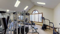 Located at 715 Meadow Wood Dr in Crescent Springs, Kentucky, this dedicated fitness room offers a thoughtfully designed space for comprehensive at-home workouts. The room is defined by its soaring vaulted ceiling, which lends an open and airy feel, while a distinctive arched window with warm wood framing serves as a prominent architectural focal point. Vertical blinds provide adjustable natural light, and skylights fitted with panel lighting illuminate the space evenly from above.

A full-length wall mirror spans one side of the room, visually expanding the space and reflecting the well-rounded collection of exercise equipment within. The fitness area accommodates a multi-station cable machine, a treadmill, a stationary air bike, an elliptical-style stepper, and a flat bench, offering both cardiovascular and strength training options. The carpeted floor provides comfort underfoot throughout.

Rich wood panel doors connect the fitness room to adjacent areas, including a casual sitting area visible through the doorway, adding context to the broader layout. An exercise guidelines board is mounted on the wall, reinforcing the room's functional, informative character. The overall ambiance is clean, practical, and purpose-built for fitness, combining architectural charm with everyday utility.