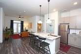 This 2-bedroom, 2-bathroom residence offers a smartly designed single-floor layout that balances comfort and functionality. The open-concept living and dining area serves as the heart of the home, featuring rich hardwood-style flooring and a ceiling fan that enhances the airy atmosphere. A generously sized kitchen overlooks the living space and is highlighted by a granite-topped breakfast bar with pendant lighting, providing both additional seating and a casual dining option. The kitchen also features a stainless steel sink and ample counter space. The living area is bathed in natural light through large windows dressed with elegant navy curtains, creating a warm and inviting ambiance. Access to the outdoors is provided through glass-panel doors off the living room. Curated wall art and lush indoor plants add a stylish, lived-in character to the shared spaces. The bedrooms are accessible directly from the main living area, offering privacy and separation from the common spaces. The overall design aesthetic is contemporary and cohesive, with neutral wall tones and modern fixtures throughout that lend a clean, polished look to every room.
