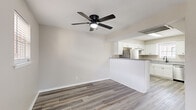 This single-floor residence offers a thoughtfully designed layout comprising a living room, kitchen, bedroom, hallway, and closet. The open-concept living area is anchored by light-toned luxury vinyl plank flooring that runs throughout the entire space, creating a cohesive and clean aesthetic. A ceiling fan with a built-in light fixture provides both comfort and illumination in the living room, which flows seamlessly into the kitchen via a peninsula-style half-wall. The kitchen is well-equipped with white shaker cabinetry, granite-look countertops, stainless steel appliances including a refrigerator and dishwasher, and a window above the sink that brings in natural light. A skylight overhead further brightens the space. The bedroom is generously sized, featuring two windows that allow ample natural light, a ceiling fan, and a built-in vanity with a large mirror and storage drawers. A sliding glass door in the bedroom provides access to the exterior. The walk-in closet is outfitted with dual hanging rods and shelving on both sides, offering substantial storage capacity. Neutral wall tones and clean white trim throughout give the space a fresh, modern feel.