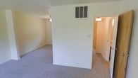 This two-story space presents a straightforward, functional interior with a neutral color palette throughout. The main living area visible in the available imagery features wall-to-wall carpeting in a muted gray tone, contributing to a quiet and understated atmosphere. Walls are finished in a clean off-white, lending a bright, open feel to the room. A central HVAC vent is positioned high on the wall, accompanied by a thermostat, indicating a climate-controlled environment. A partial dividing wall creates a subtle separation within the living space, adding a degree of structural definition without fully closing off the area. An adjacent room, accessible through a doorway, continues the carpeted flooring and similarly light-toned walls, suggesting consistent finishes across the level. Ceiling-mounted lighting provides overhead illumination in both visible areas. The overall design is minimal and practical, prioritizing livability over ornate detailing. With two floors, the space offers room for functional separation between living and private areas, making it a versatile layout suitable for a variety of uses.